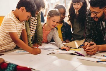 Un docente con gafas guía a un grupo diverso de niños que trabajan con papeles y cuadernos alrededor de una mesa, en un espacio luminoso de aprendizaje colaborativo.