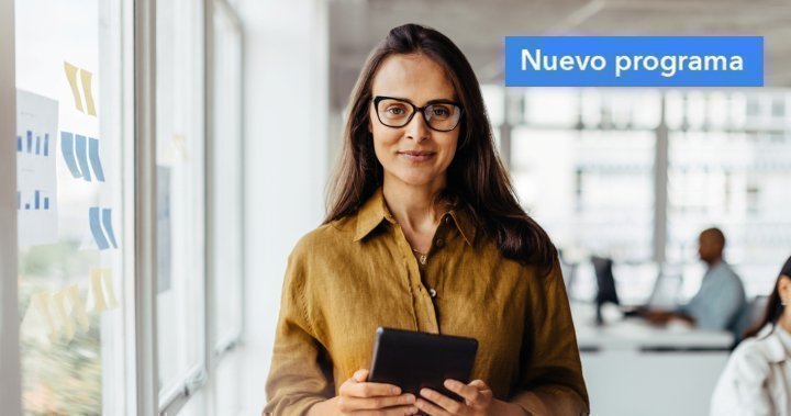 Mujer con gafas sostiene una tablet en una oficina luminosa; cartel ‘Nuevo programa’ en la esquina