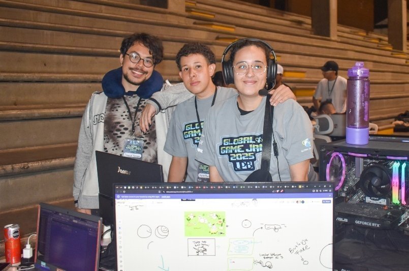 Grupo de jóvenes en un puesto de videojuegos, con camisetas grises y lanyards, trabajando frente a computadoras y monitor en un evento.