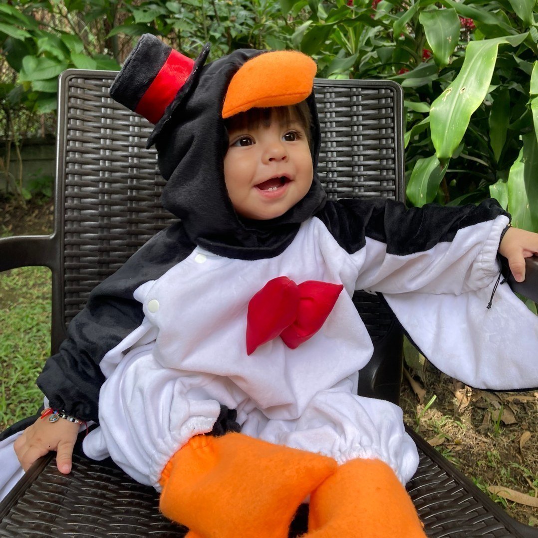 Una bebé sonriendo vestida con un disfraz de pingüino blanco y negro, con pico naranja, sentado en una silla de jardín al aire libre.